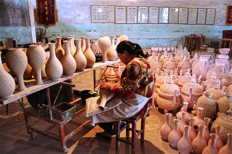 In dieser Fabrik in Xian, die inzwischen nicht mehr existiert, trägt eine Arbeiterin Schnitt– und Ritzdekore auf. Sie ist von Dutzenden Gefäßen umgeben, teils sehr großen. An der Wand im Hintergrund hängen zahlreiche Urkunden und Auszeichnungen.
