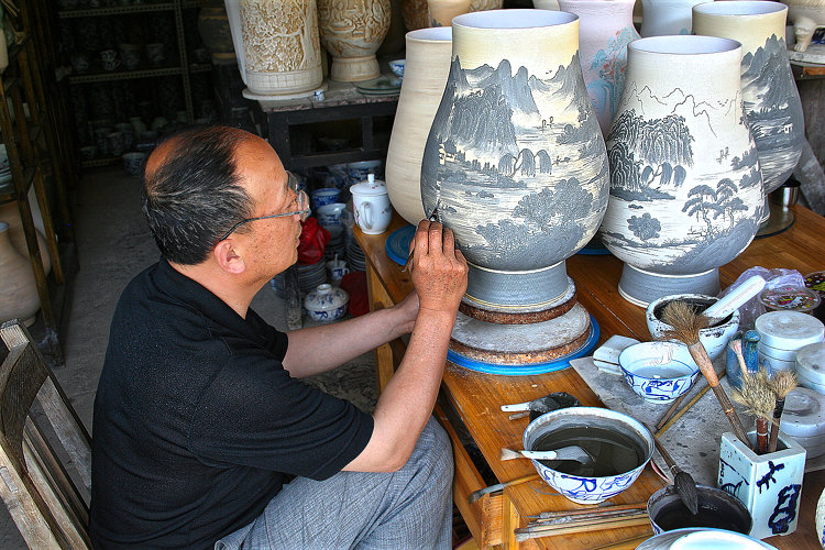 Ein Porzellanmaler trägt Landschaftsbilder auf die unglasierten Gefäße auf.