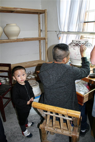 Ein Porzellanmaler in seiner privaten Werkstatt mit seinem kleinen Sohn, der fragend in Kamera schaut.