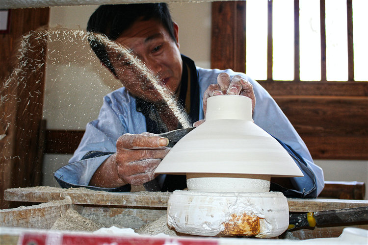 Abdreher in Jingdezhen bei der Arbeit. Die Geschwindigkeit der Abdrehscheibe ist offenbar hoch, denn die fortgeschleuderten Tonspäne fliegen in einem stattlichen Bogen.